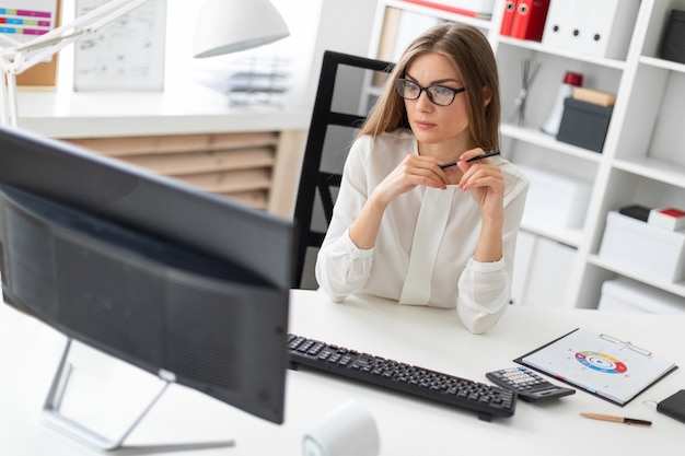 Una niña sentada en el escritorio de la oficina, con un lápiz en la mano y mirando el monitor.