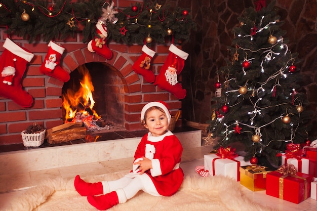 Niña sentada junto a la chimenea y el árbol de navidad con cajas de regalo