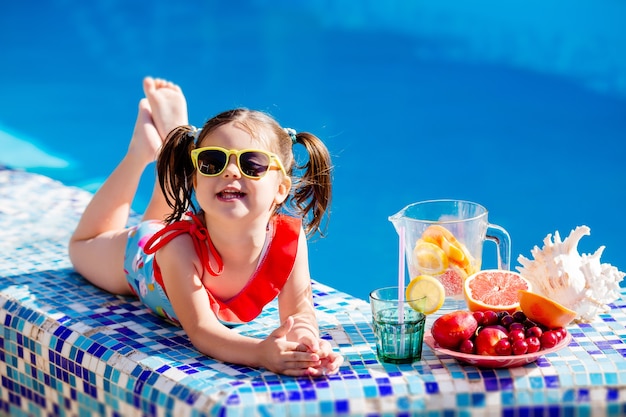 Niña sentada junto a la piscina bebiendo limonada