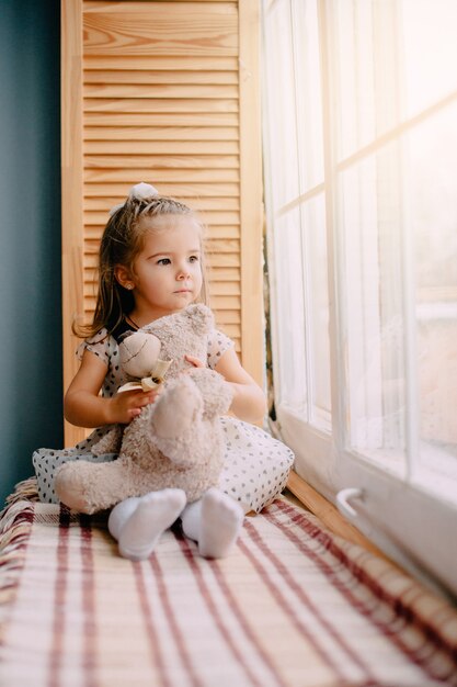 Niña sentada junto a la ventana con oso de juguete