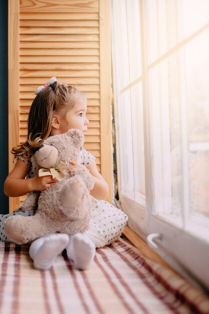 Niña sentada junto a la ventana con oso de juguete