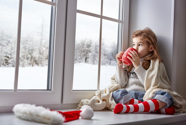 Niña sentada junto a la ventana con una taza de bebida caliente y mirando el bosque de invierno