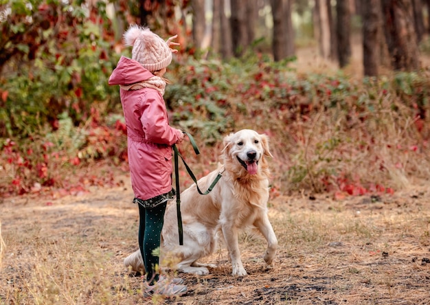 Niña sentada de lado sosteniendo la correa de golden retriever en las manos en el bosque de otoño