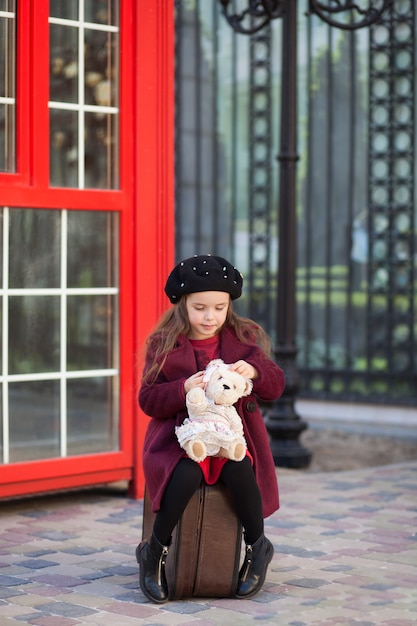 La niña está sentada en una maleta con un oso de peluche. cabina telefónica roja de londres. primavera. otoño. viaje. viaje. londres, inglaterra. chica de escuela. vacaciones escolares. niña en una boina y abrigo. de viaje