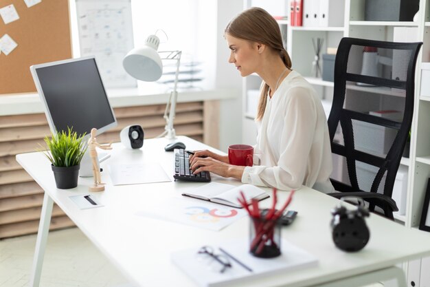 Una niña sentada en una mesa en la oficina, sosteniendo una taza roja en la mano y trabajando en la computadora.