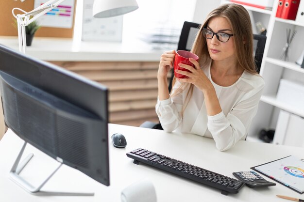 Una niña sentada en una mesa en la oficina, sostiene una taza roja en la mano y mira el monitor.