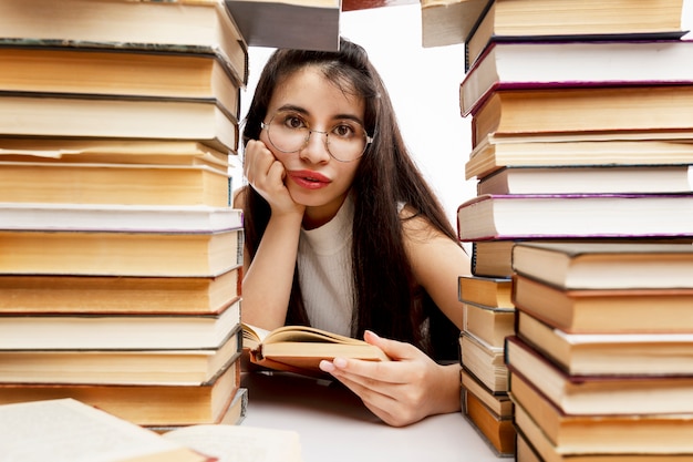 Niña sentada en una mesa con una pila de libros. Triste morena con gafas. Entrenamiento y educación. De cerca.