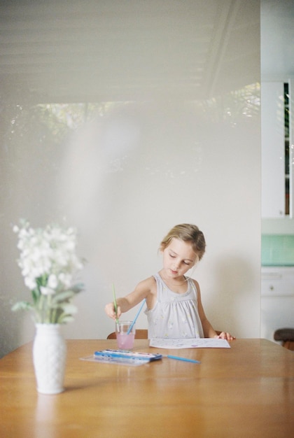 Niña sentada en una mesa pintando un jarrón con flores blancas