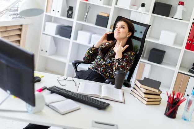 Una niña sentada en la oficina en la computadora Escritorio y hablando por teléfono.