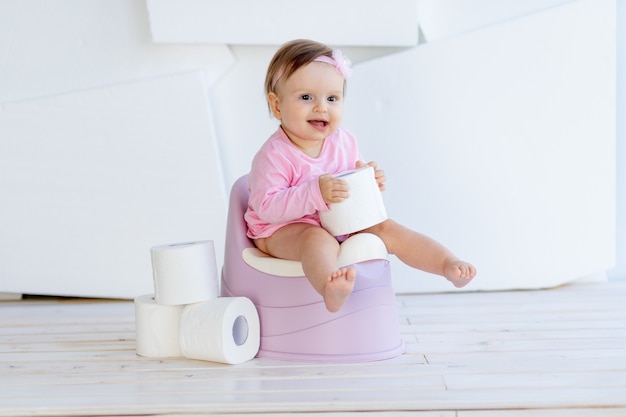 Una niña sentada en un orinal con ropa rosa en una habitación luminosa y juega con papel higiénico