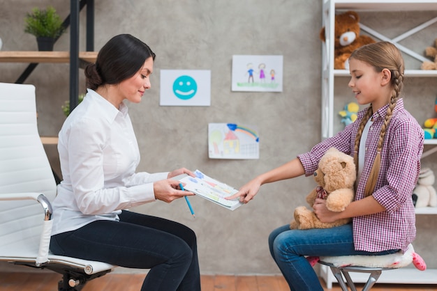 Foto niña sentada con un oso de peluche apuntando al papel de dibujo que muestra su psicóloga