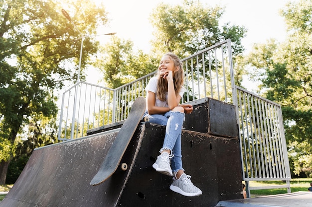 Niña sentada con patineta en la rampa deportiva Equipo deportivo para niños Adolescente activo con patineta en el patio de recreo del parque de patinaje