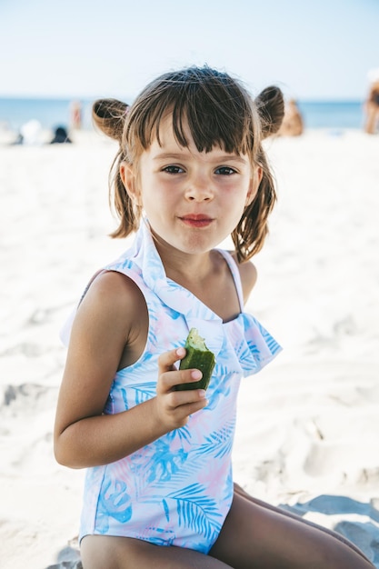 Niña sentada en la playa a la sombra y comiendo un pepino