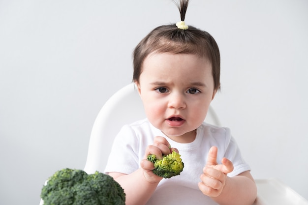 Niña sentada en una silla de bebé comiendo brócoli.