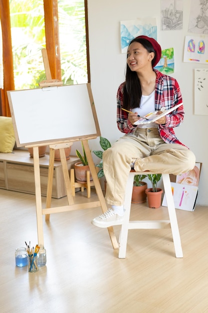 Niña sentada en una silla con caballete para dibujar paleta de colores y pincel en la habitación