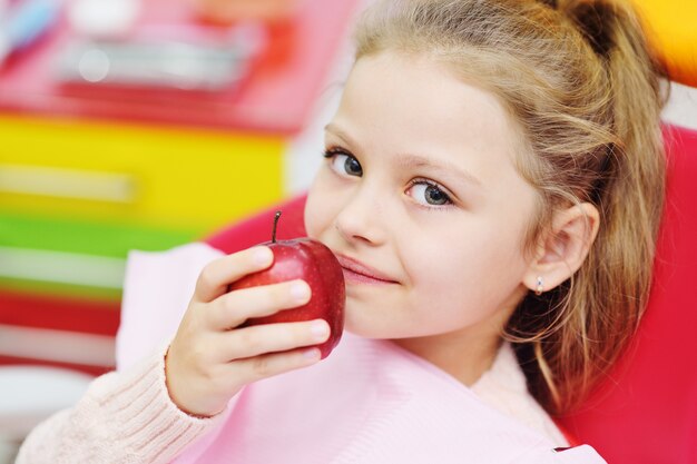 Niña sentada en una silla dental roja sonriendo con una manzana roja en sus manos. Odontología pediátrica, dientes de leche.