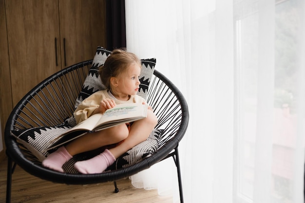 Niña sentada en una silla junto a la ventana leyendo un libro