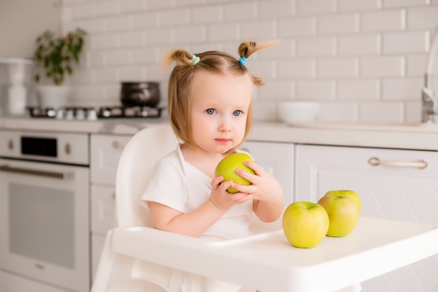 Niña sentada en la silla de un niño en la cocina comiendo manzanas verdes