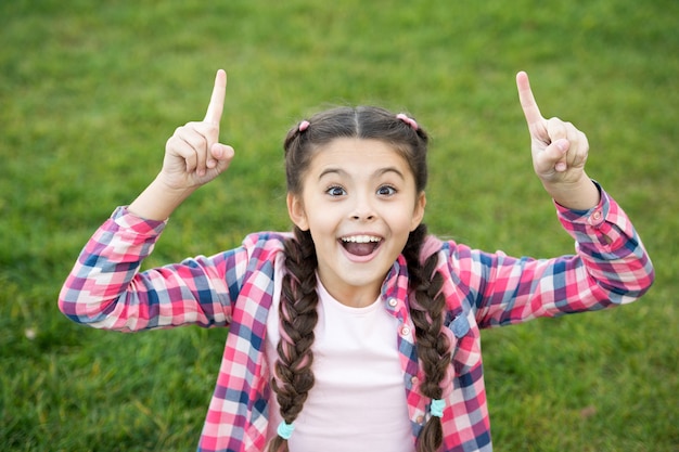 Niña sentada sobre la hierba verde brillante. Pequeña belleza mostrando sus dedos. Expresión feliz en la cara. Sonrisa bonita. Sensación de emoción y alegría. Bonita jovencita divirtiéndose. Tener idea.