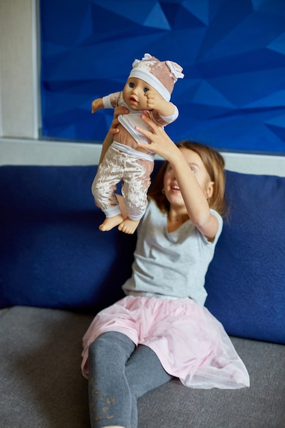 Niña sentada en el sofá de la habitación en casa jugando con la muñeca, cantando canciones, como mamá.