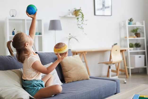 Niña sentada en el sofá y jugando con modelos de planetas en la sala de estar de casa