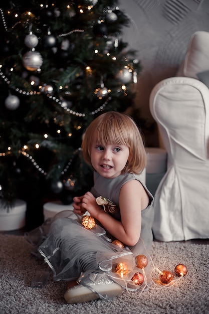 Niña sentada en el suelo junto a un árbol de navidad y sosteniendo una guirnalda en sus manos