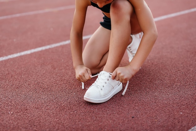 Una niña se sentó para atarse los cordones de los zapatos antes de entrenar en el estadio durante la puesta de sol Deportes infantiles y un estilo de vida saludable