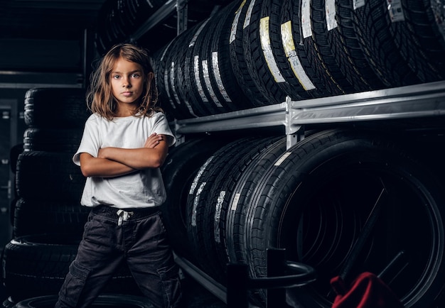 Una niña seria está parada con las manos cruzadas cerca de un estante con neumáticos en un almacén oscuro.