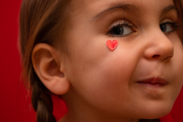 Una niña seria mira a la cámara con el corazón en la mejilla con coletas en un fondo rojo Foto de alta calidad