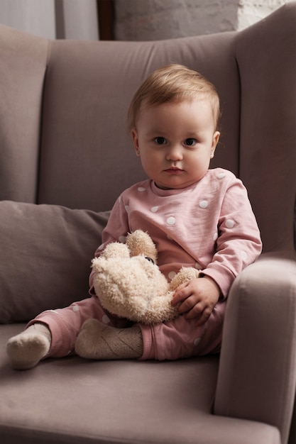 Una niña seria en pijama rosa con lunares blancos se sienta en un sillón gris y sostiene un juguete en sus manos.