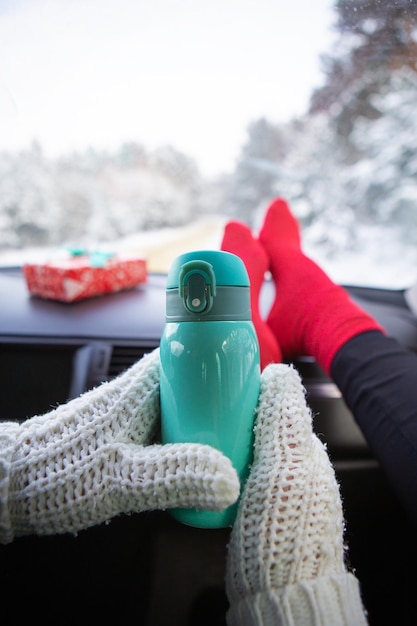 Una niña se sienta en calcetines rojos en un automóvil en invierno y se calienta con café de un termo contra el fondo de un bosque invernal Conceptos de viaje e invierno