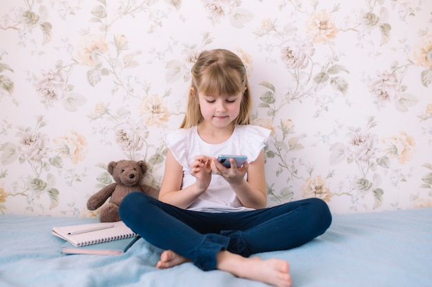 Una niña se sienta en la cama en el elegante dormitorio, sostiene el teléfono y lee algo en el teléfono inteligente. Concepto de comunicación