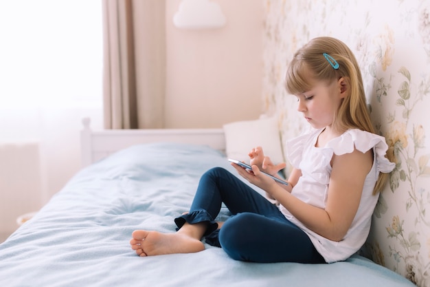 Una niña se sienta en la cama en el elegante dormitorio, sostiene el teléfono y lee algo en el teléfono inteligente. Concepto de comunicación