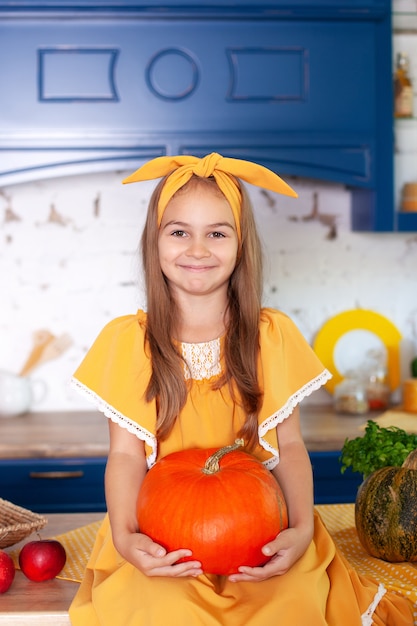 Niña se sienta en la mesa en la cocina tiene gran calabaza.