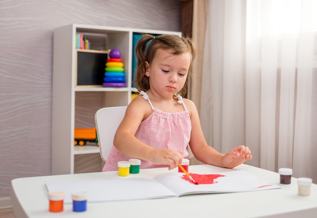 Una niña se sienta en una mesa y dibuja en la mesa con un pincel y pinturas