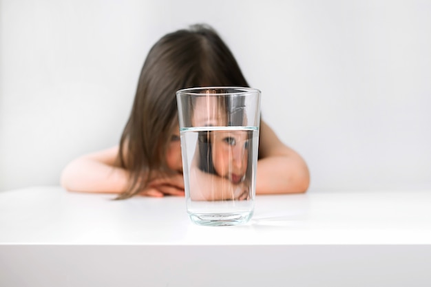 La niña se sienta en la mesa molesta. La niña está triste porque no quiere beber agua.