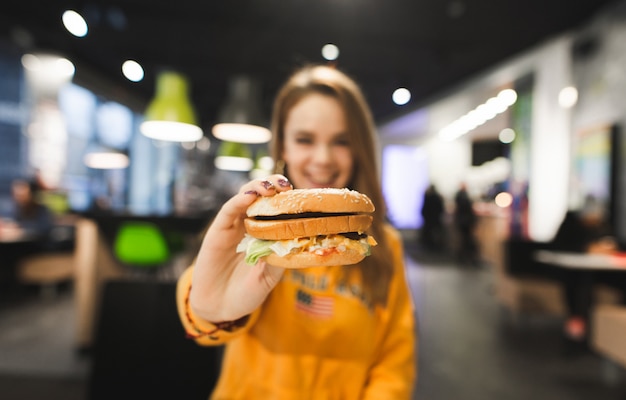 La niña se sienta a la mesa en un restaurante de comida rápida y muestra una gran hamburguesa apetitosa a la cámara