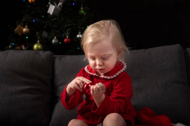 La niña se sienta en un sofá contra el fondo de un árbol de Navidad y mira un juguete de árbol de Navidad Nochevieja