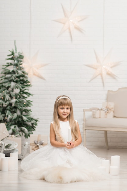 Una niña se sienta y sostiene una vela en la mano en una habitación cerca del árbol de Navidad.