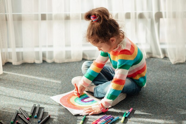 Una niña se sienta en el suelo bajo los rayos del sol y dibuja un arco iris con marcadores de colores