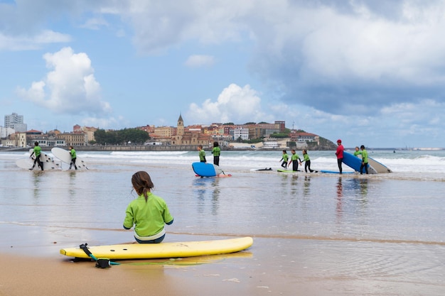 Una niña se sienta en una tabla de surf en una playa con otras personas en el fondo.