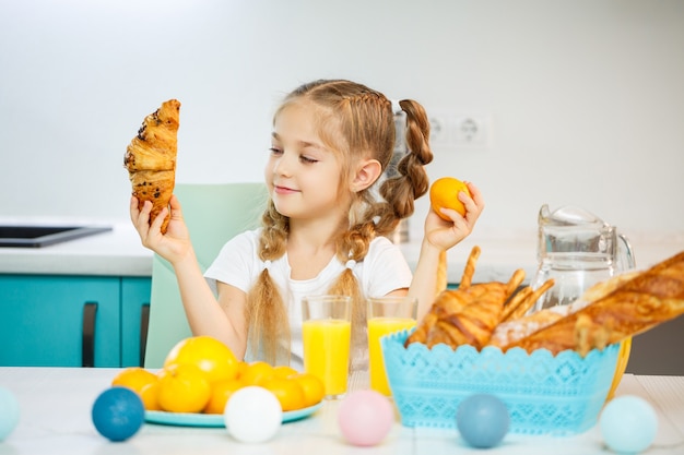 Una niña de siete años, vestida con una camiseta blanca, se sienta a la mesa de la cocina. Contiene mandarinas y un croissant recién horneado con chocolate.