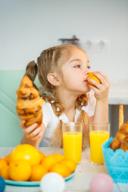 Una niña de siete años, vestida con una camiseta blanca, se sienta a la mesa de la cocina. Contiene mandarinas y un croissant recién horneado con chocolate.