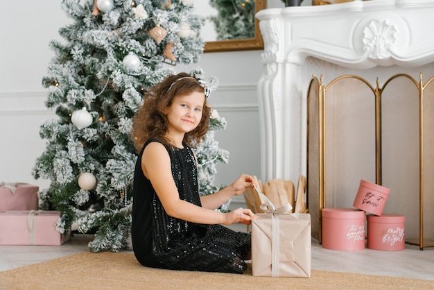 Una niña de siete años con un vestido negro con lentejuelas se sienta cerca de la chimenea en el árbol de Navidad y abre un regalo de Año Nuevo.