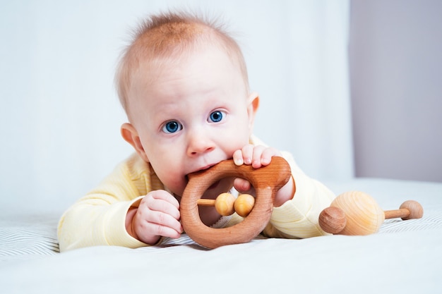 Una niña de siete meses con ojos azules yace boca abajo en una habitación luminosa y muerde un juguete de madera.