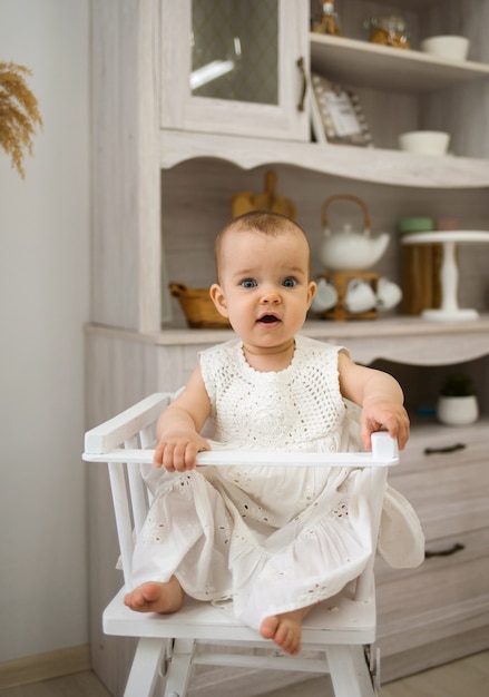 Niña en una silla de bebé blanca se sienta en la cocina y mira a la cámara