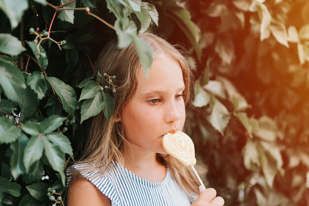 Una niña sincera y pensativa de ocho años sosteniendo piruletas y lamiendo comiendo dulces de azúcar de frutas en el fondo de un muro de cobertura de plantas verdes en una bengala al aire libre de vacaciones de verano