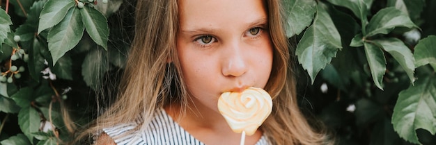 Una niña sincera y pensativa de ocho años sosteniendo piruletas y lamiendo comiendo dulces de azúcar de frutas en el fondo de un muro de cobertura de plantas verdes en una pancarta al aire libre de vacaciones de verano