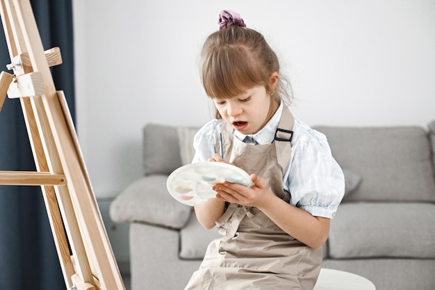 Niña con síndrome de Down con delantal beige pintando en un caballete