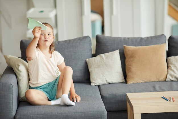 Niña con síndrome de down sentada en el sofá y jugando con avión de papel en la sala de estar